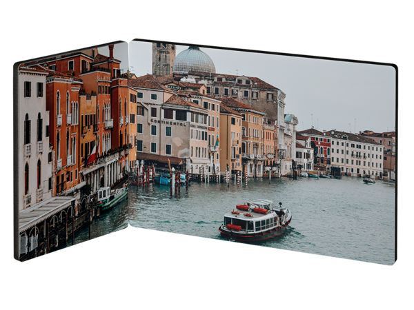 Ein zweiteiliger Fotoaufsteller zeigt ein belebtes Stadtbild am Wasser. Bunte Gebäude säumen einen breiten Kanal, auf dem mehrere Boote unterwegs sind. Im Hintergrund ist ein großer Dom mit einer hellen Kuppel zu sehen. 